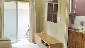 A room with a sliding glass door leading to a balcony, a small window, and a kitchenette with wooden cabinets.