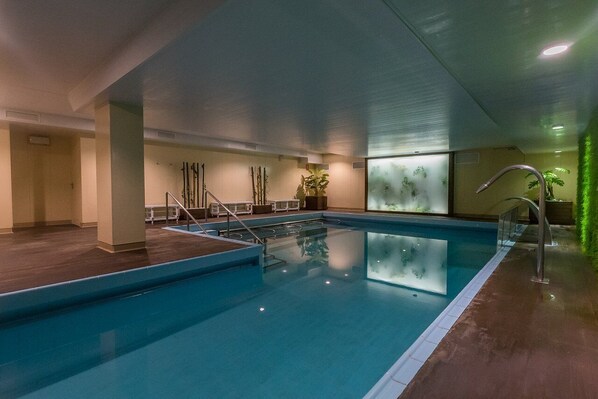 Une piscine intérieure à l’eau claire, une terrasse en bois et un mur de verre agrémenté de plantes.