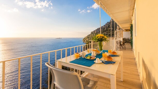 A balcony with a table set for breakfast overlooking the sea, a chair, and a white railing.