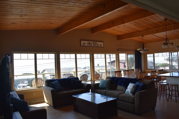 A spacious living room with large windows, a wooden ceiling, and a sign that reads "Life is better at the beach".