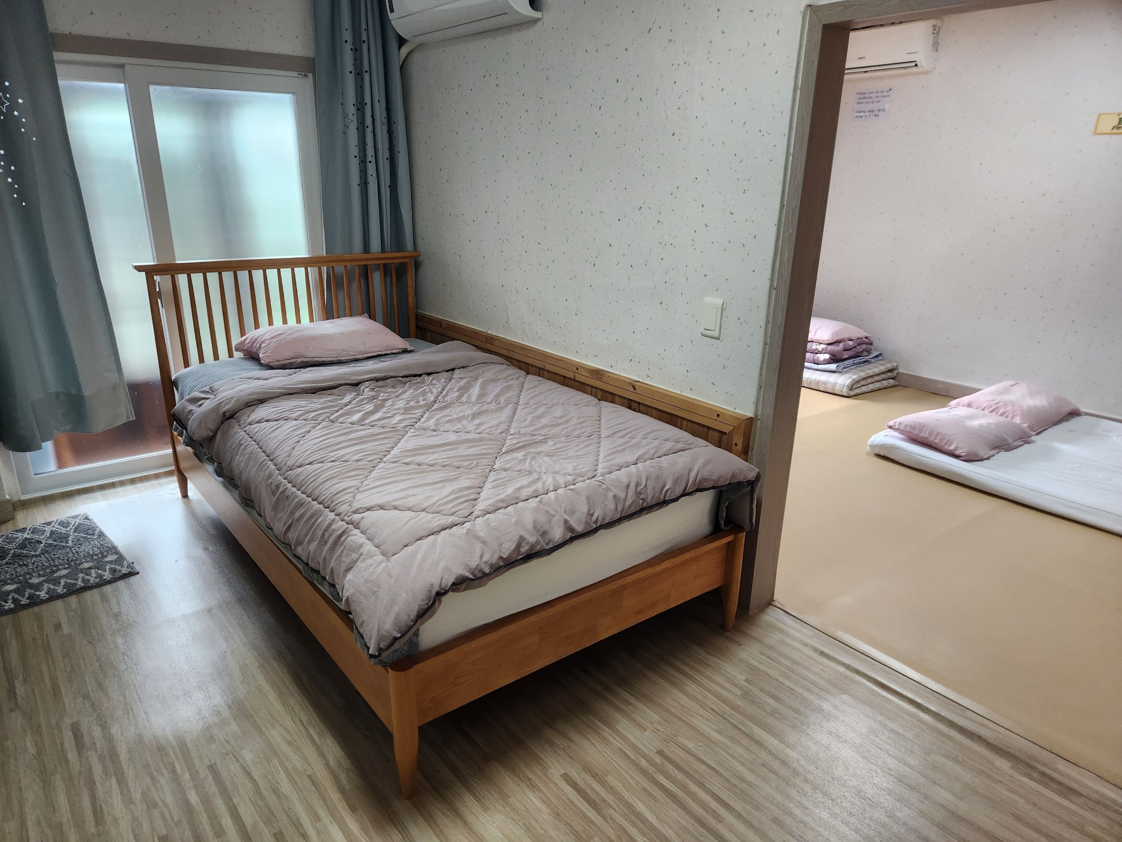 A room with a wooden bed, a small bedside table, a window with curtains, and a wall-mounted air conditioner.