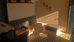 A compact living space with a sofa, a small table, and a bench. There are baskets on a shelf, a wall-mounted shelf, and a door leading outside. - Comfortable studio in the Gulf of St Tropez (Cavalaire-sur-Mer)