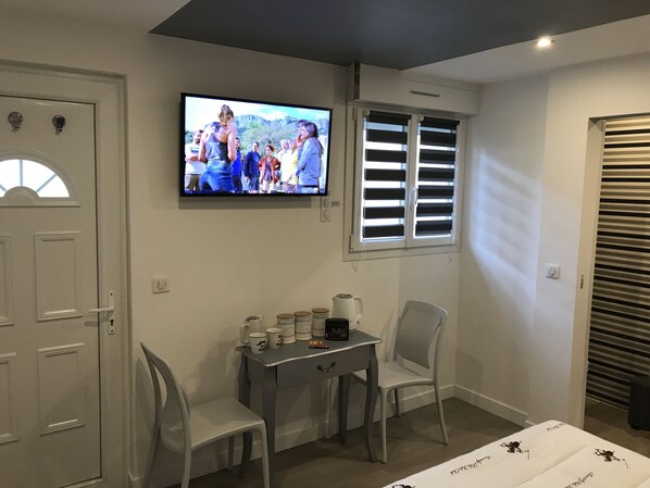 A hotel room with a television mounted on the wall, a desk with chairs, and a window with blinds.