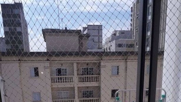 A view through a window of a building with a metal fence, a balcony, and a door with a green handle. - Apartamento Itaim Bibi (São Paulo)