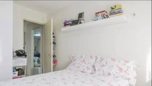 A bedroom with a bed, a white wall, and a shelf with books and a laptop. - Apartamento Itaim Bibi (São Paulo)