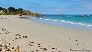 Een strand met helderblauw water, rotsformaties en enkele gebouwen op de achtergrond.