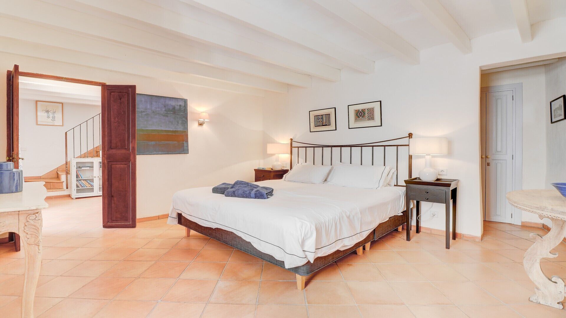 A bedroom with a large bed, wooden headboard, two bedside tables with lamps, a wooden door, and a staircase.