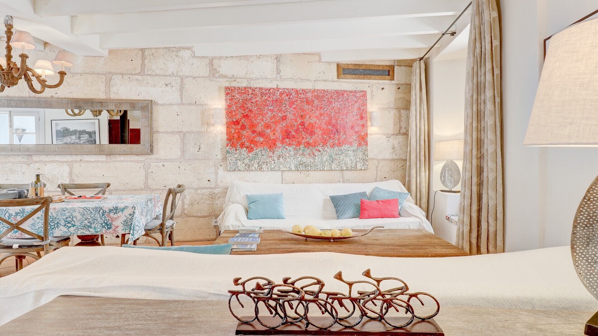 A cozy room with a rustic stone wall, a bed with colorful pillows, a dining area with a table and chairs, and a chandelier.