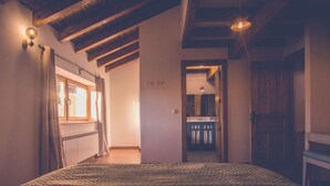 A bedroom with a large bed, wooden beams, a window with curtains, and a view into the bathroom.