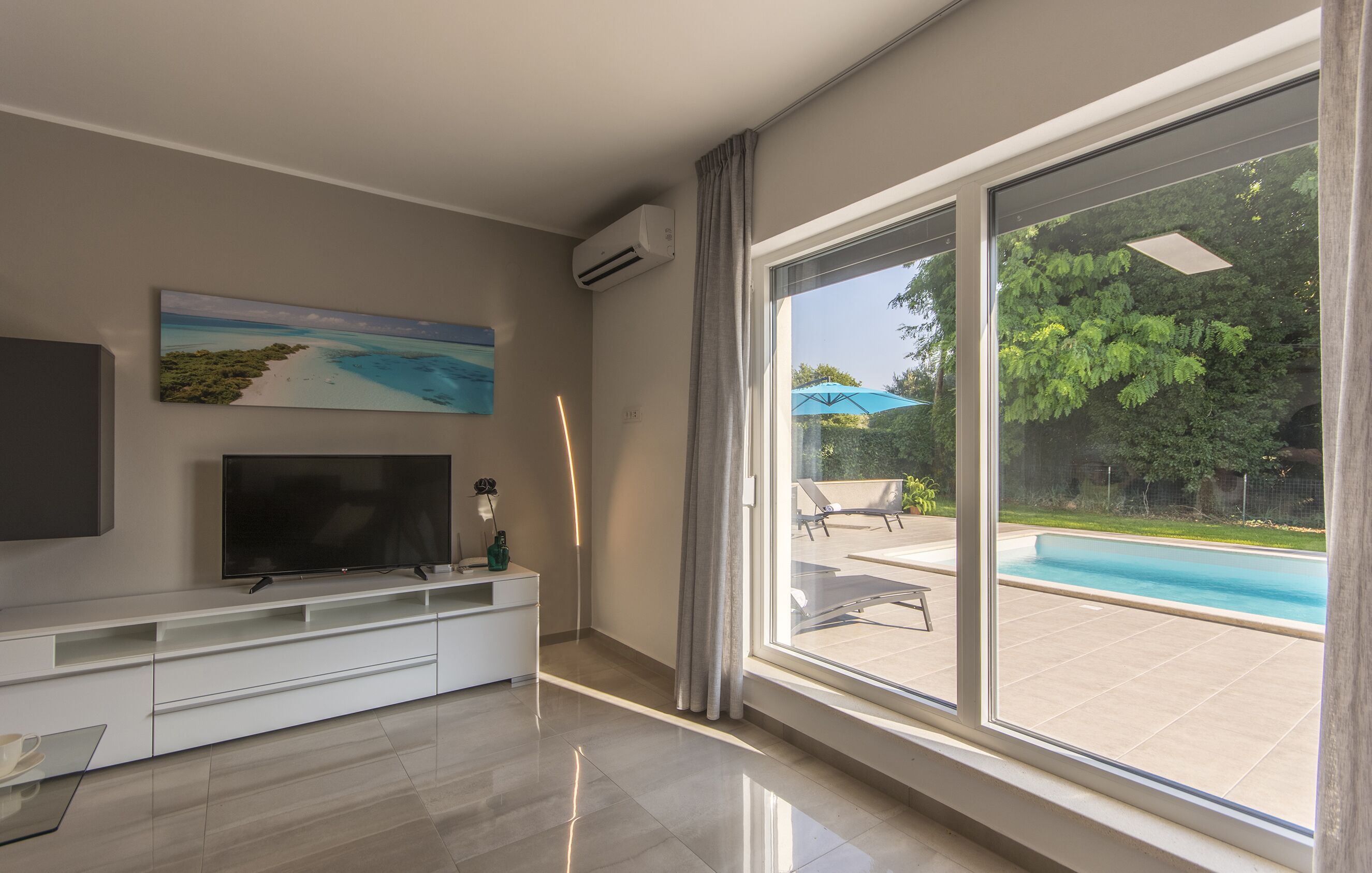 Ein modernes Wohnzimmer mit einem Flachbildfernseher, einem Gemälde vom Strand und einer großen Glasschiebetür, die zu einem Poolbereich führt.