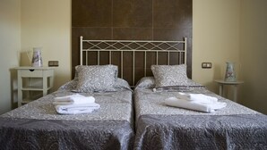 Een tweepersoonsbed met beddengoed met een patroon en twee kussens, een klein nachtkastje met een vaas, en een lichtschakelaar aan de muur.