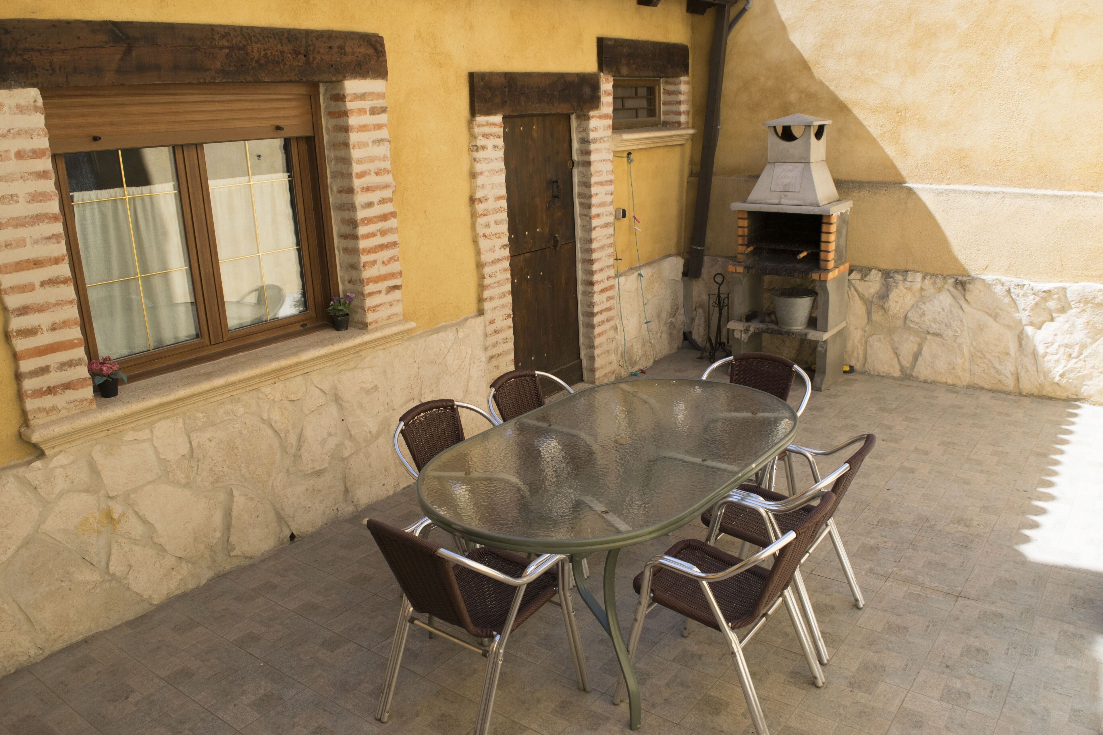 Un patio al aire libre con una mesa de vidrio rodeada de sillas de mimbre, un muro de ladrillo y un suelo de piedra.