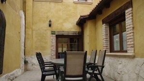 Een terras buiten met een ronde tafel en zes stoelen, een deur en een raam, met een gevel van baksteen.