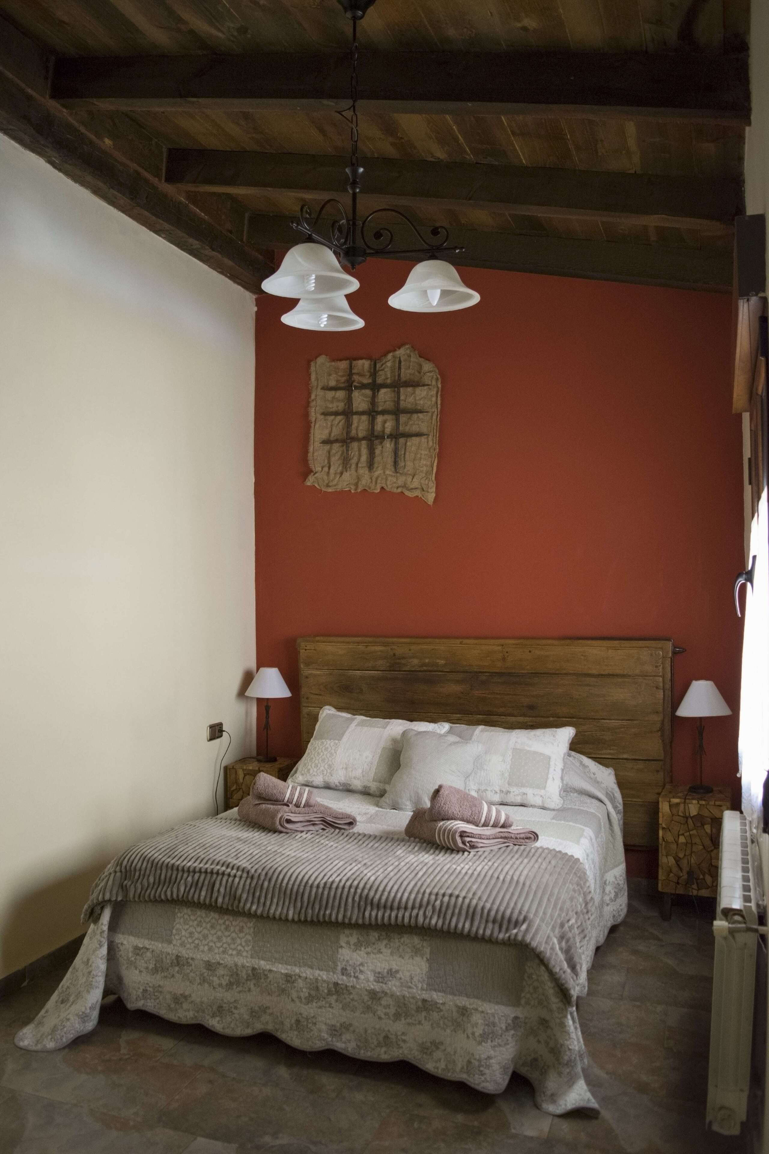 A bedroom with a wooden bed, two bedside lamps, a red wall, and a rustic ceiling.