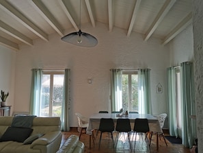 Uma sala de estar ampla com área de jantar, paredes brancas e teto alto com vigas aparentes.