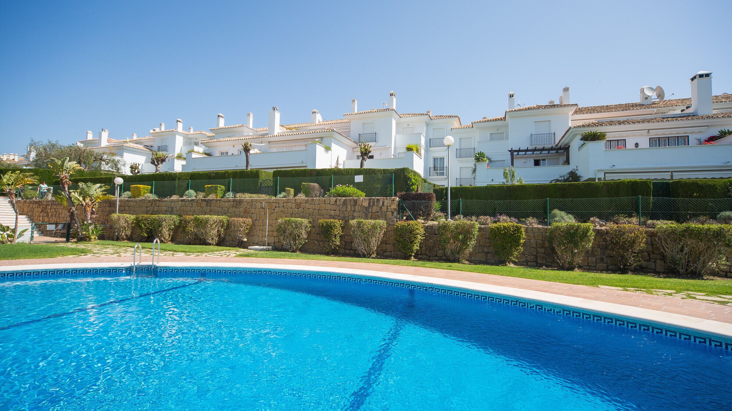 A swimming pool with a clear blue water, surrounded by a stone wall and a well-maintained hedge. In the background, there are white buildings with red-tiled roofs and balconies.