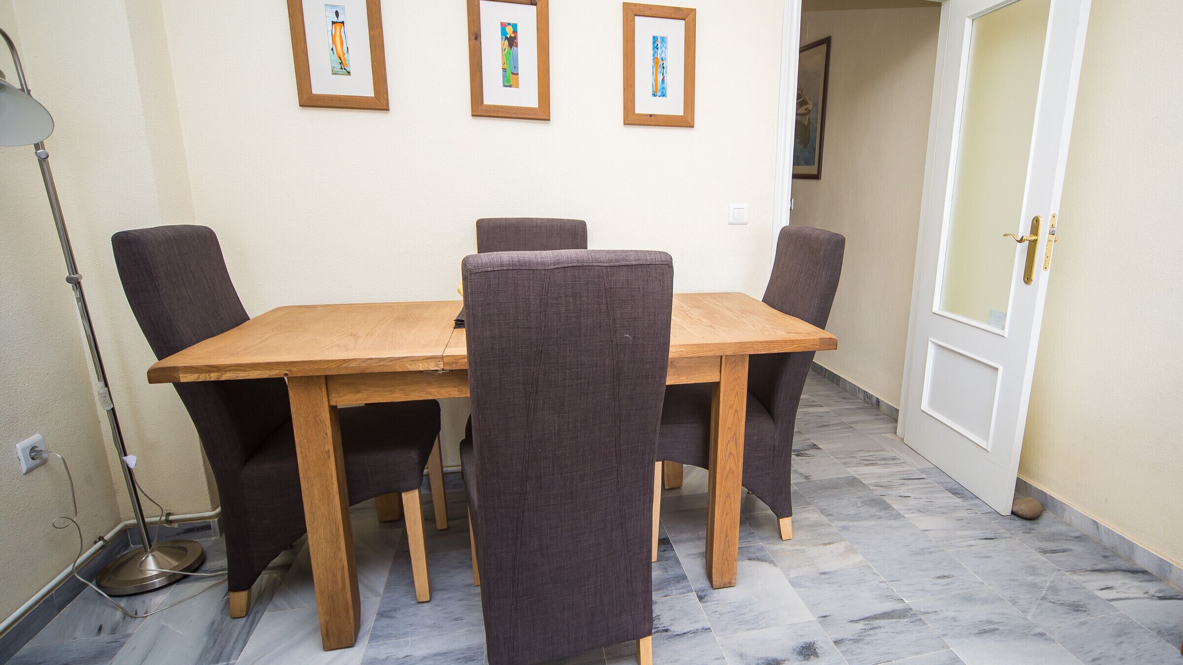 Un comedor con una mesa de madera y cuatro sillas, una lámpara de pie y tres obras de arte enmarcadas en la pared.