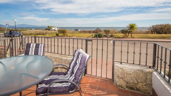 En terrasse med et glasbord og stribede stole, en stenvæg og udsigt til havet og bjergene.
