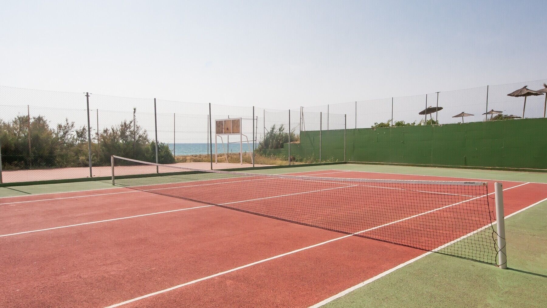 Ein Tennisplatz mit Netz, umgeben von einem grünen Zaun und Blick auf das Meer.