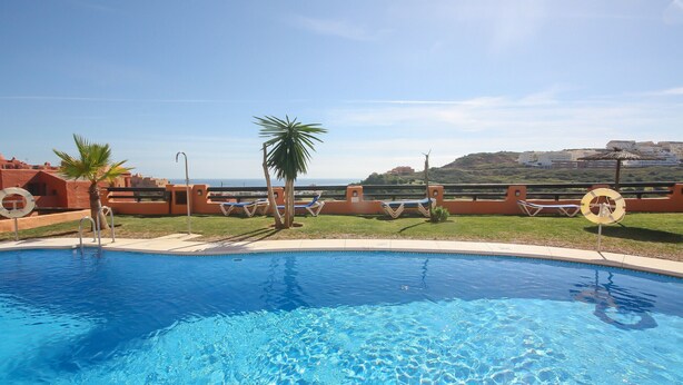 A swimming pool with clear blue water, surrounded by a low wall with lounge chairs and a palm tree. - Coto Real Andros 201 (Manilva)