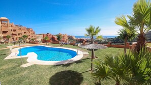 A swimming pool surrounded by grass, palm trees, and a view of buildings in the background. - Coto Real Andros 201 (Manilva)