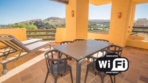 A balcony with a table and chairs, a sun lounger, and a view of a hilly landscape. - Coto Real Andros 201 (Manilva)