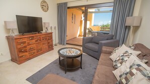 A living room with a TV, wooden dresser, round clock, grey sofa, and a view to the outside. - Coto Real Andros 201 (Manilva)