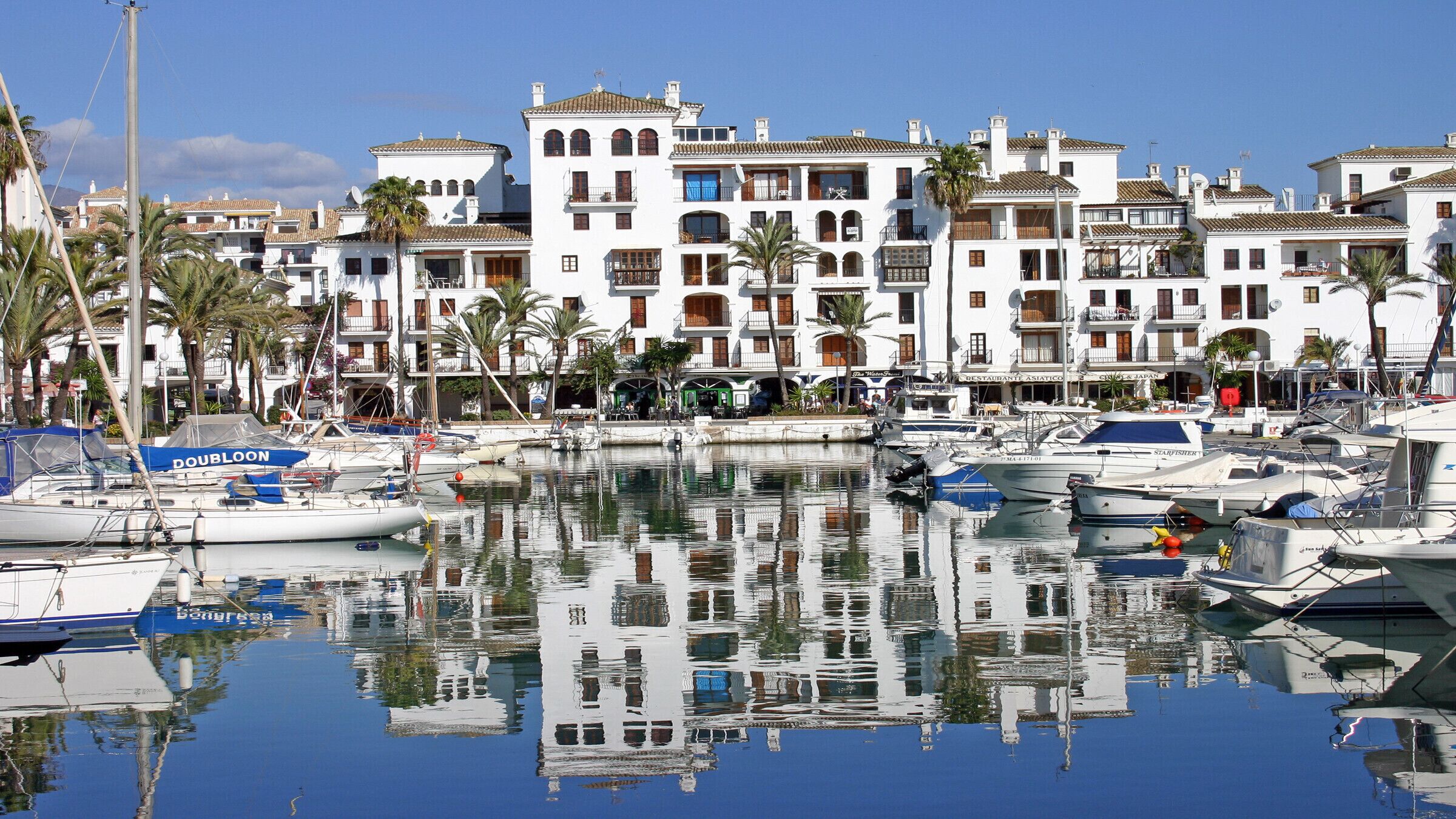 Un puerto deportivo con barcos amarrados, rodeado de edificios blancos con balcones y palmeras.