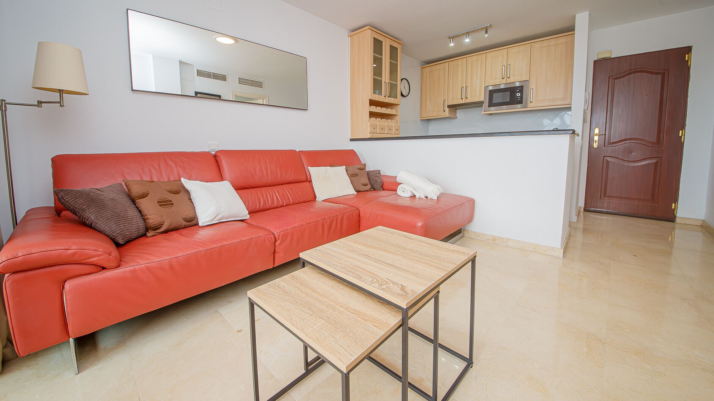 A living room with a red sofa, a small wooden table, and a kitchen area with wooden cabinets and a microwave.