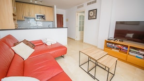 Ein Wohnzimmer mit rotem Sofa, einem kleinen Holztisch, einem Fernseher auf einem Holzschrank und einer Küchenzeile mit Holzschränken und einer Mikrowelle.