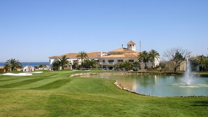 Un parcours de golf avec un étang et une fontaine, entouré de palmiers et d’un grand bâtiment doté d’une tour.