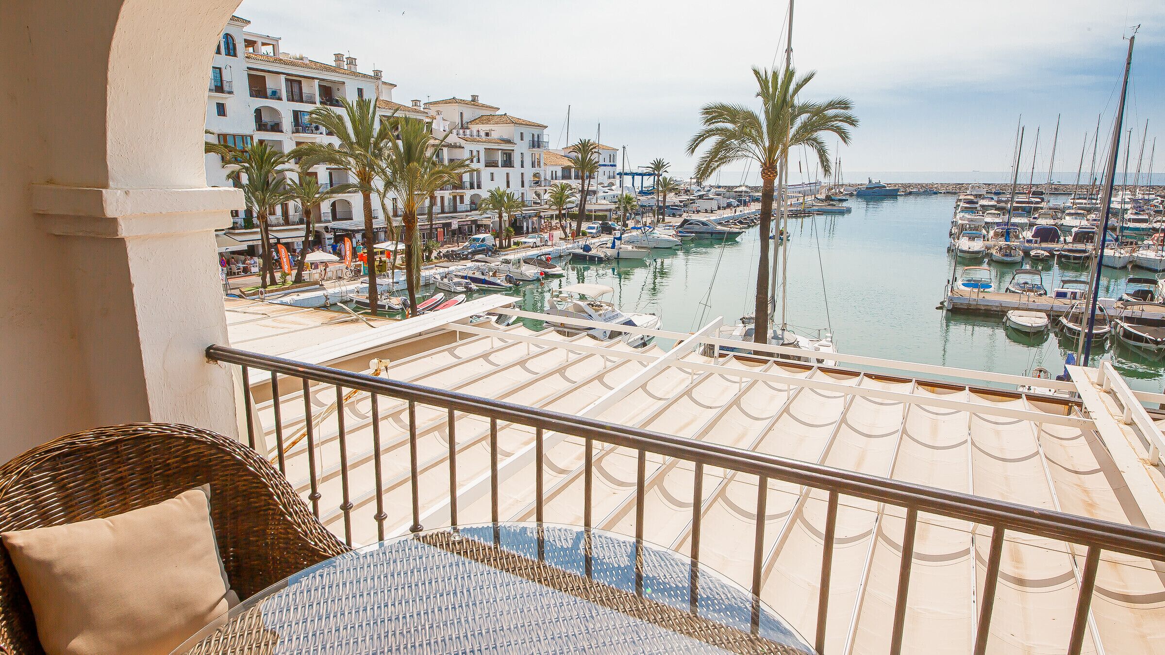 A balcony with wicker furniture, overlooking a marina with boats and buildings.