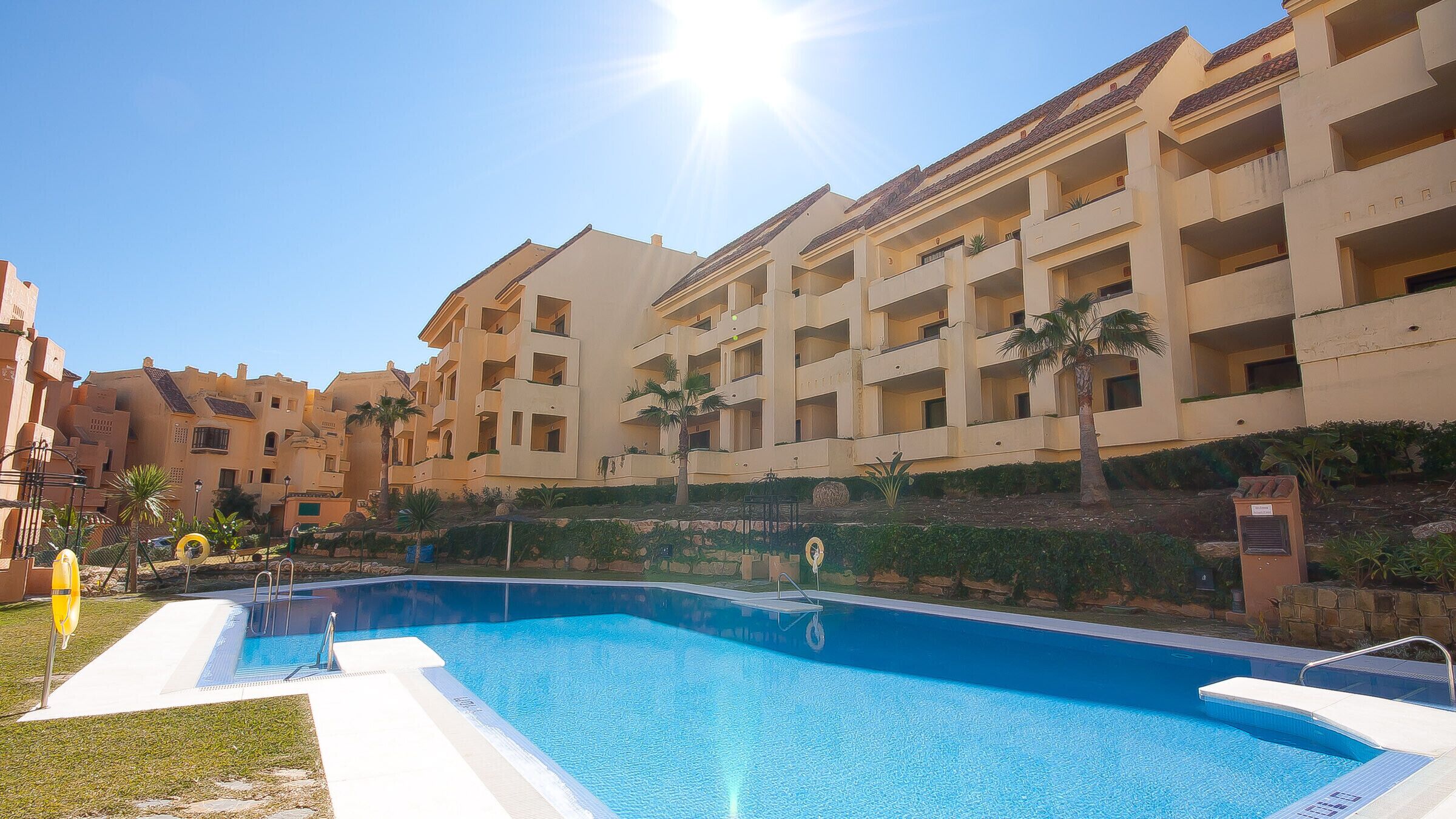 Une piscine à l’eau d’un bleu limpide, entourée d’une pelouse bien entretenue et bordée d’une rangée d’immeubles à plusieurs étages dotés de balcons.
