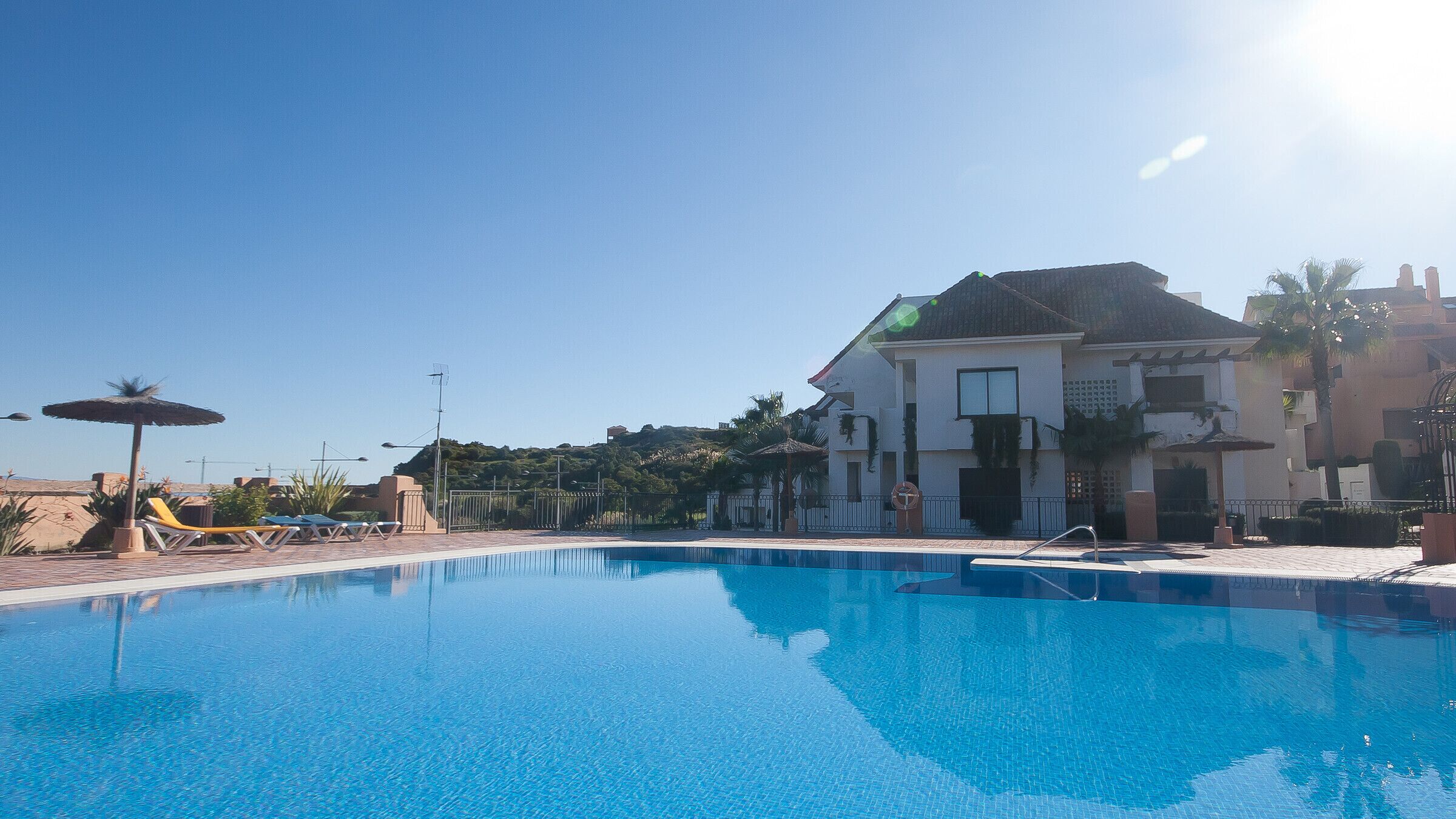 A swimming pool with lounge chairs and umbrellas, surrounded by a building with a tiled roof and a view of a hilly landscape.