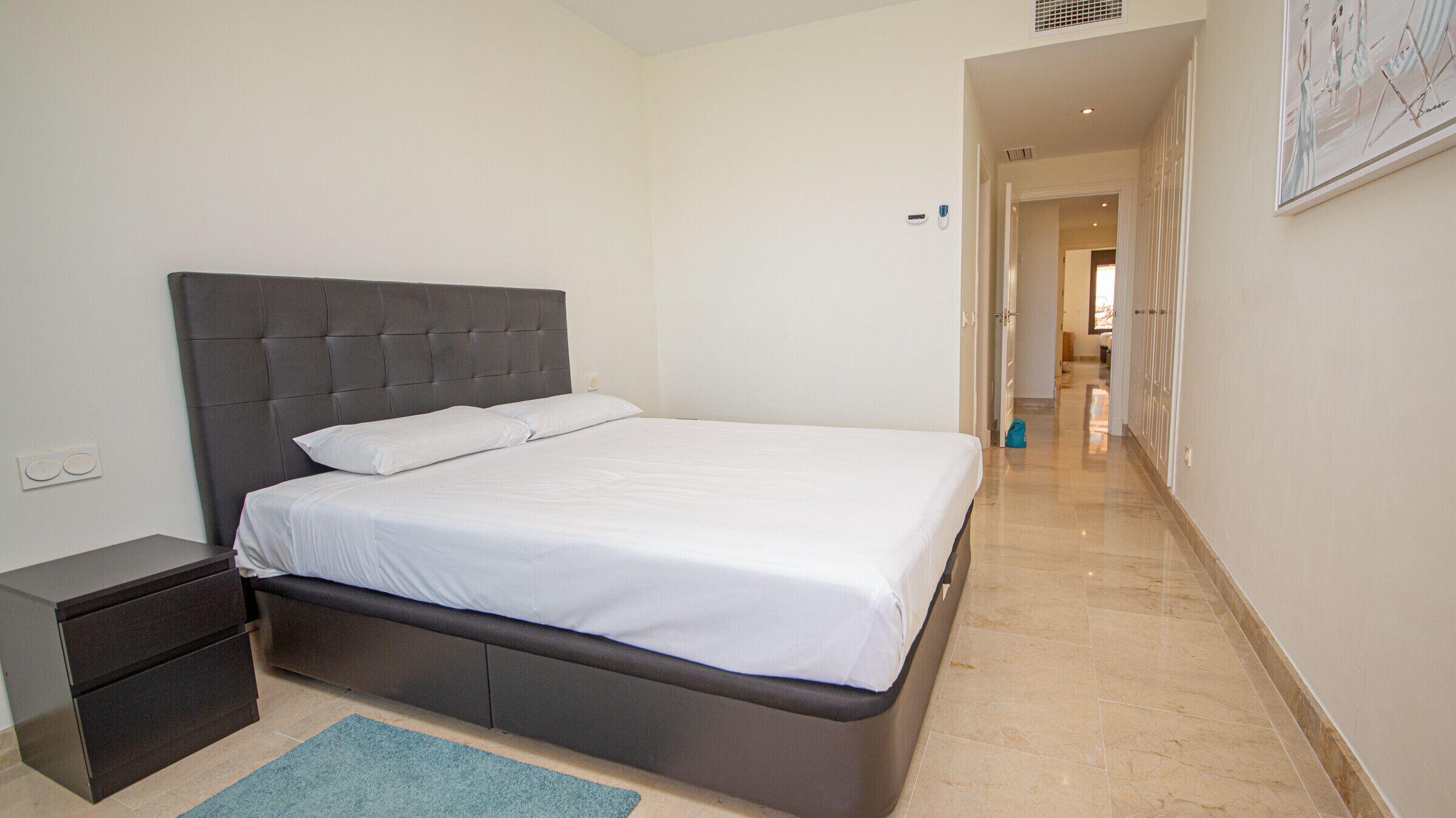 A neatly made bed in a hotel room with a dark headboard, a small bedside table, and a blue rug.