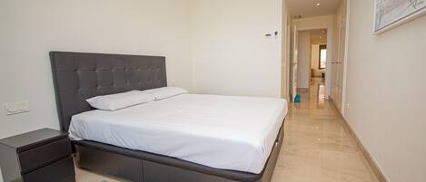 A neatly made bed in a hotel room with a dark headboard, a small bedside table, and a blue rug.