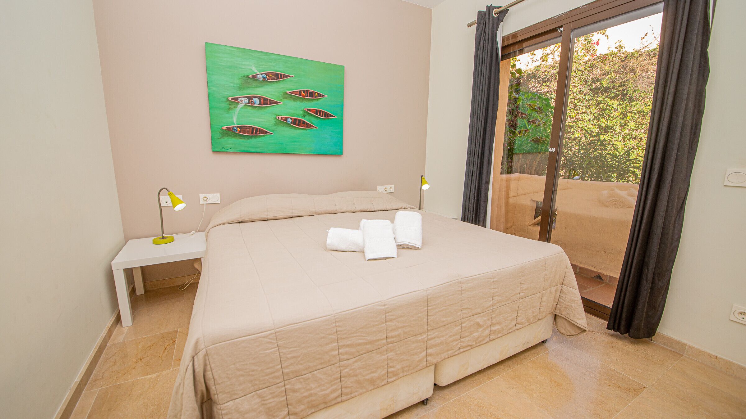 A bedroom with a bed, a nightstand, a lamp, a painting, and a view through a glass door.