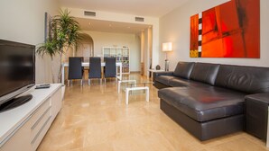 A modern living room with a flat-screen TV, a leather sofa, a glass coffee table, and a dining area with chairs and a table. - Coto Real 102 Khios. (Manilva)