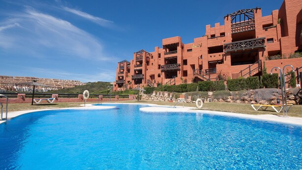 A swimming pool with lounge chairs and a red brick building in the background. - Coto Real 102 Khios. (Manilva)