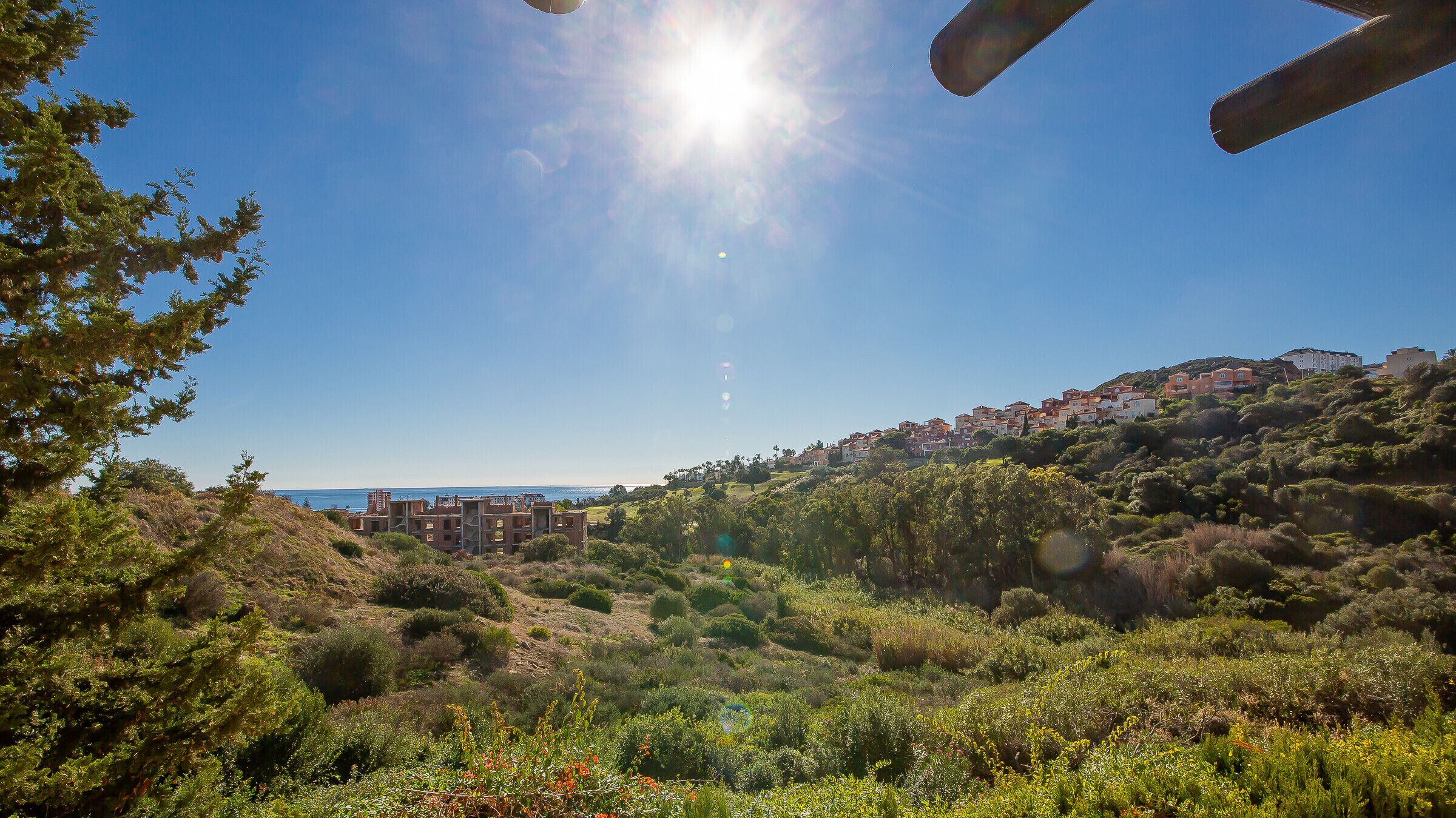 A coastal landscape with a clear sky, dense greenery, and a distant view of buildings on a hillside.