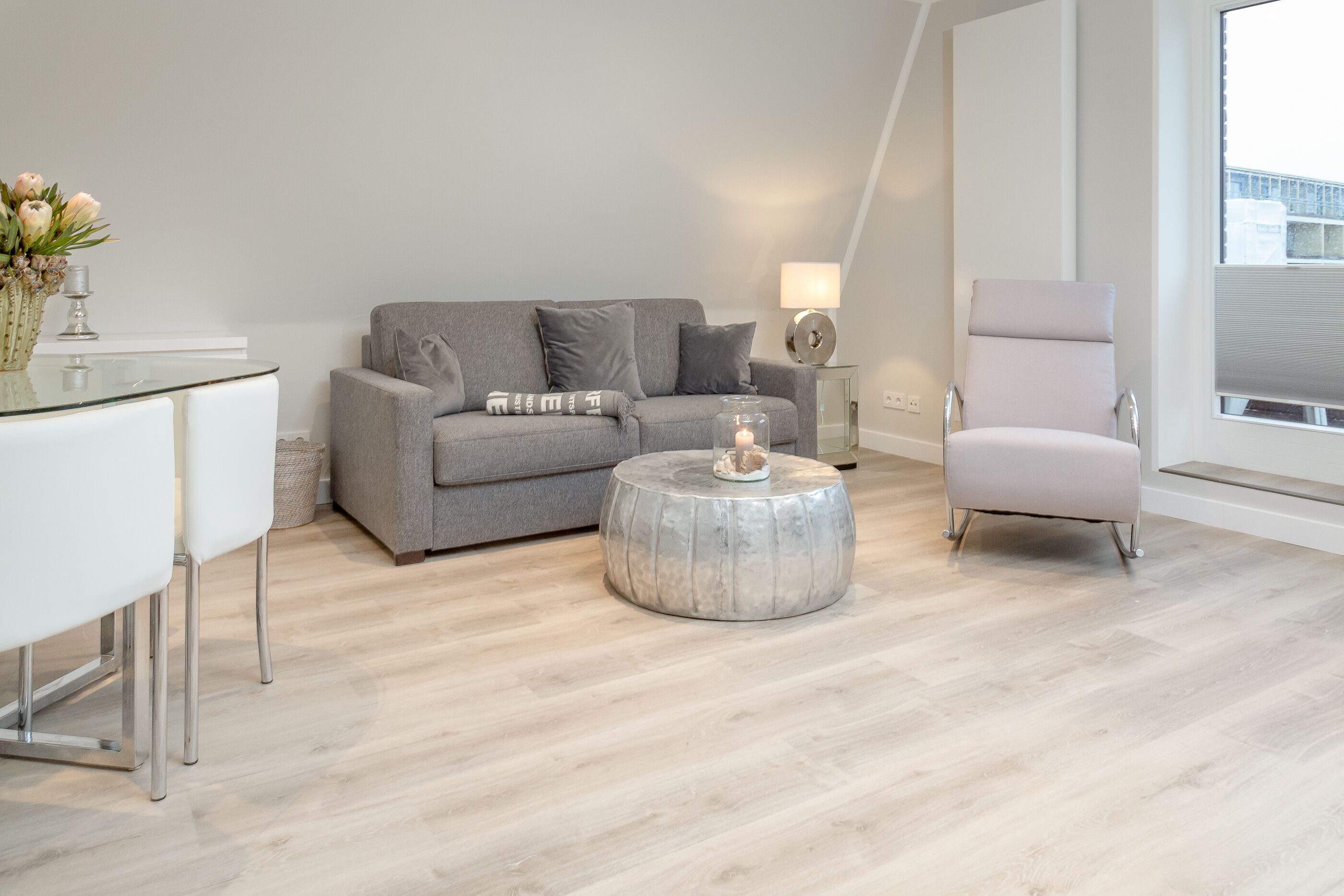 A modern living room with a grey sofa, a round coffee table, a white dining table, and a tall chair.