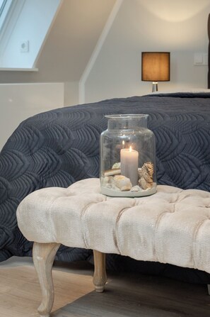 A glass jar with a candle and seashells on a cushioned bench in a room with a dark quilted bedspread and a lamp on the wall. - Penthouse maisonette Lagmöwe (Westerland)