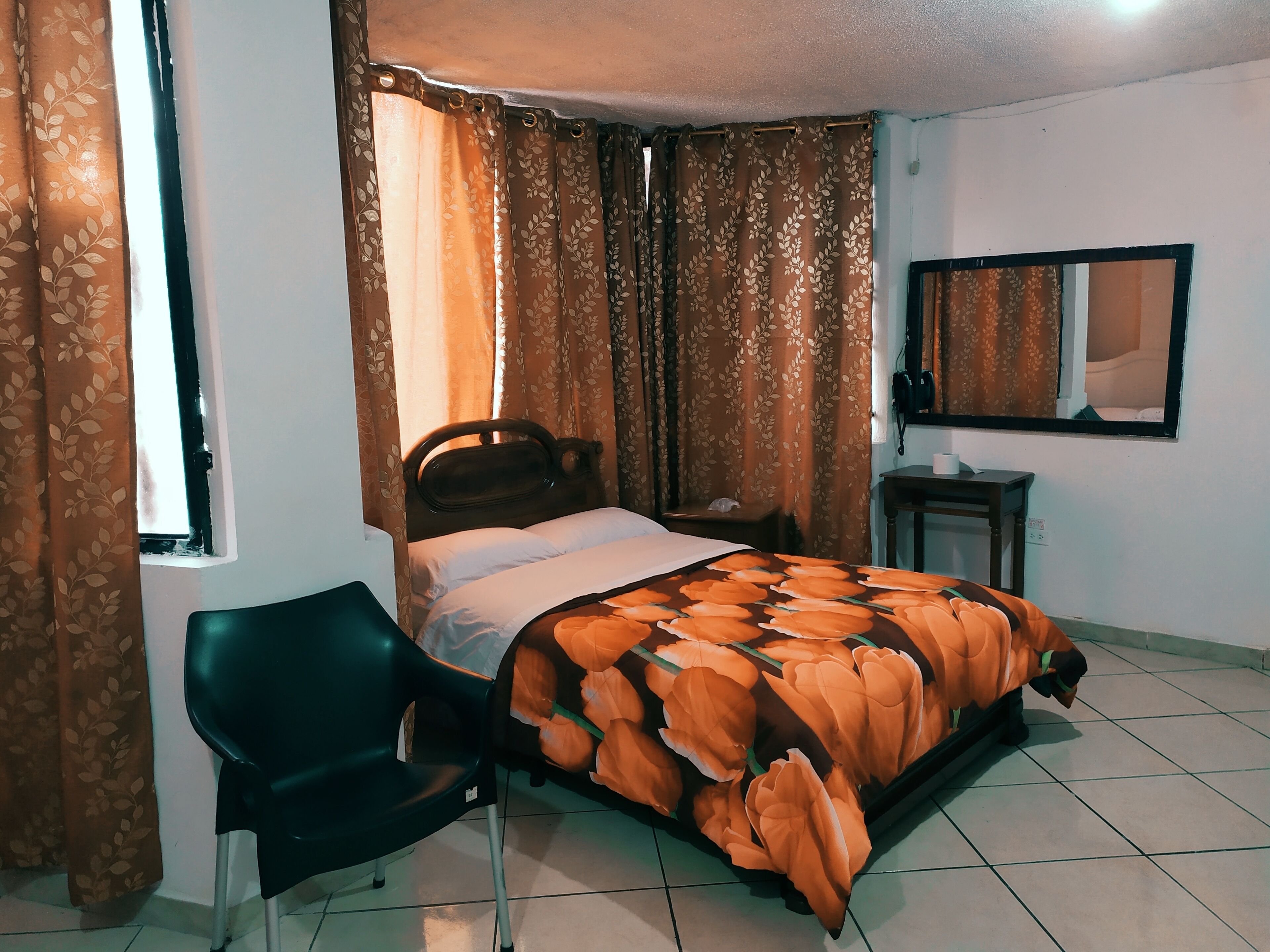 Un dormitorio con una cama, una silla, una mesita de noche, un espejo y cortinas.