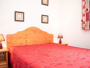 A room with a wooden headboard bed, a red patterned bedspread, two bedside lamps, and two framed pictures on the wall. - Cozy 2-room + balcony for 6 in Vignec (Vignec)