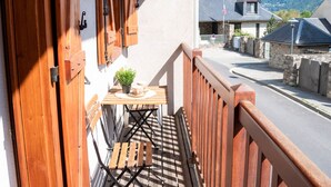 Ein Balkon mit Holzgeländern, einem kleinen Tisch mit einem Blumentopf und Blick auf einen Berg sowie Gebäude im Hintergrund.
