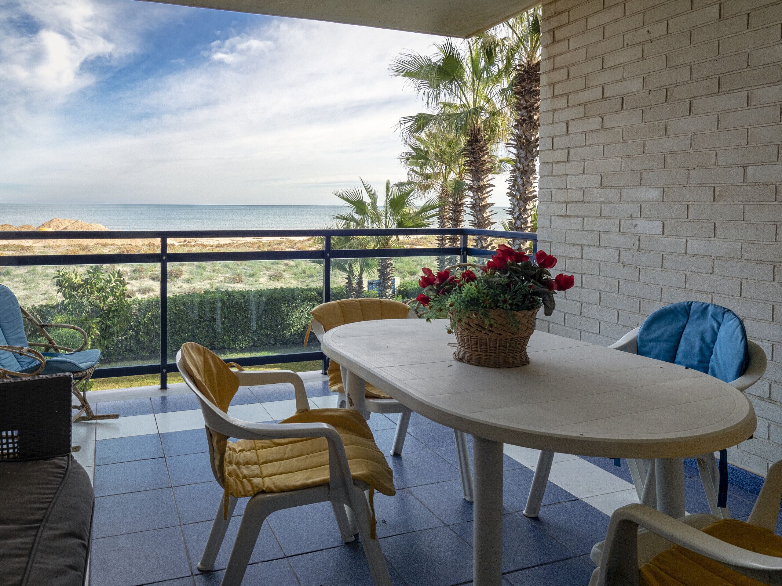 A balcony with a table and chairs, a wicker basket with red flowers, and a view of the sea and palm trees.