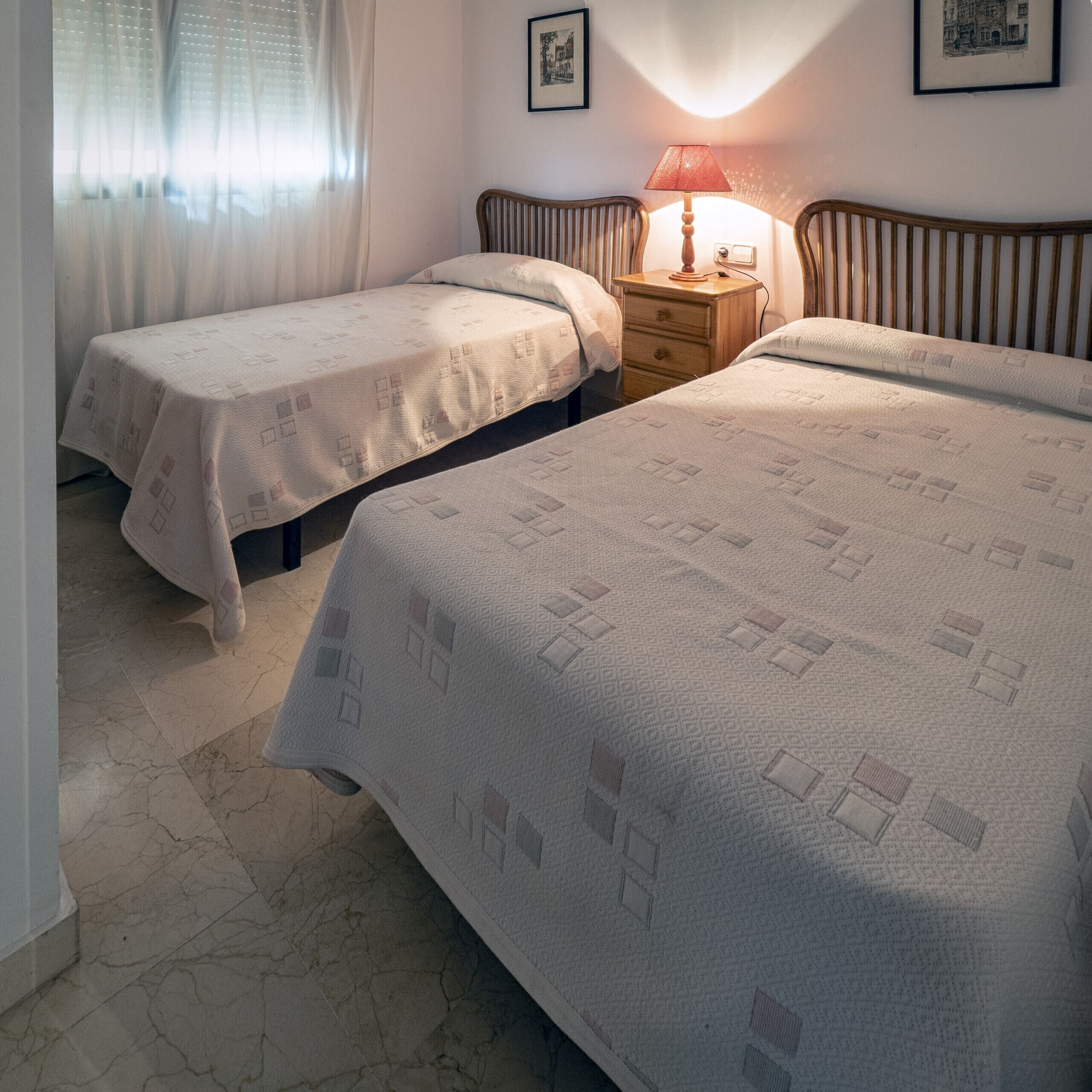 A bedroom with two beds, a nightstand, a lamp, and a window with curtains.