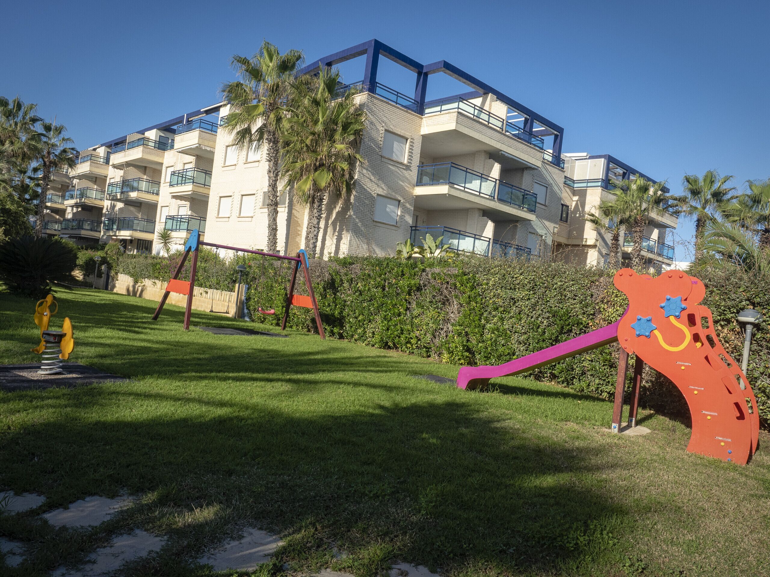 A modern apartment building with balconies, a playground with swings and a slide, and palm trees in the background.