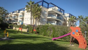 A modern apartment building with balconies, a playground with swings and a slide, and palm trees in the background. - Dorasal Bl. A esc. IV 1º Solo Familias! (Xeraco Playa)