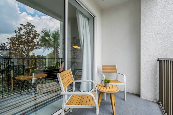 A balcony with two white chairs, a small round table with a plant, and a glass door leading to a patio area with a striped rug. - IS1-208 - 3BR condo sleeps 8 near universal (Orlando)
